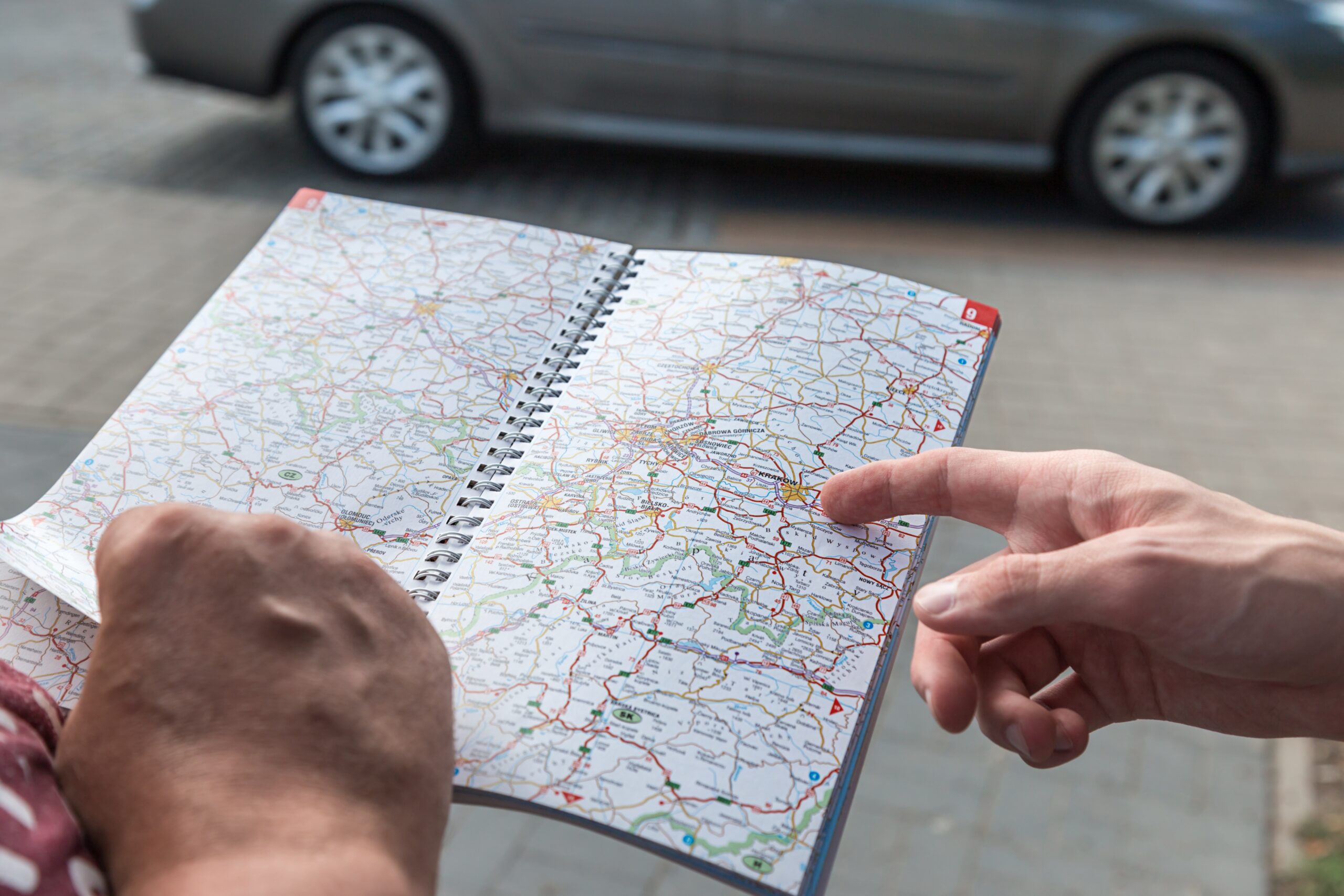 Eine Person hat einen Straßenatlas aufgeschlagen und eine weitere Person zeigt mit einem Finger die beste Route. Im Hintergrunfd ist ein Auto zu sehen.