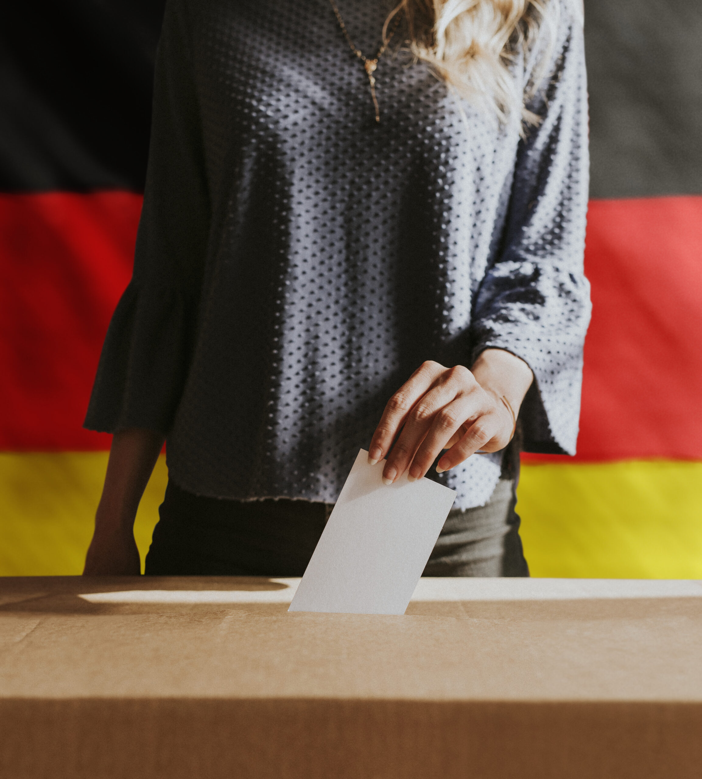 Eine Frau steckt einen verschlossenen Wahlumschlag in eine Wahlurne. Man sieht nur den Oberkörper und die Arme der Frau. Im Hintergrund befindet sich die deutsche Flagge (schwarz-rot-gold).