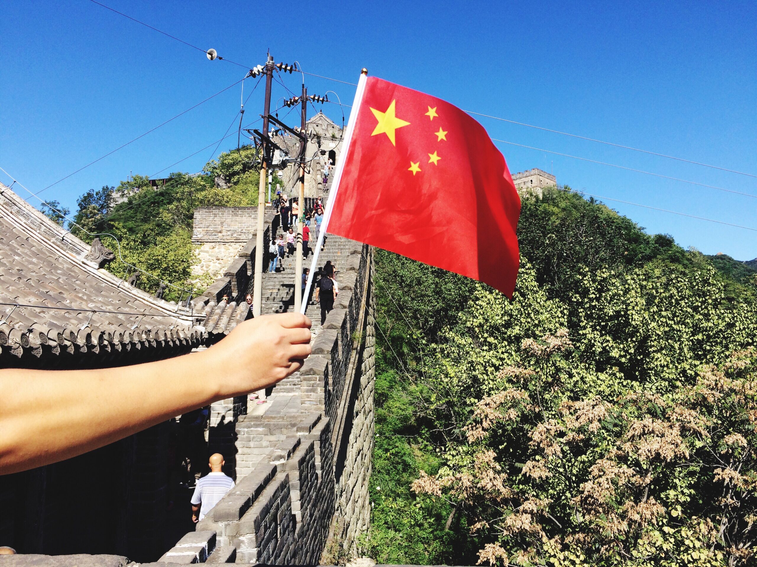 Eine Person hält eine chinesische Flagge in der Hand. Von der Person ist nur der Arm und die Hand zu sehen. Die Person geht auf der chinesischen Mauer.