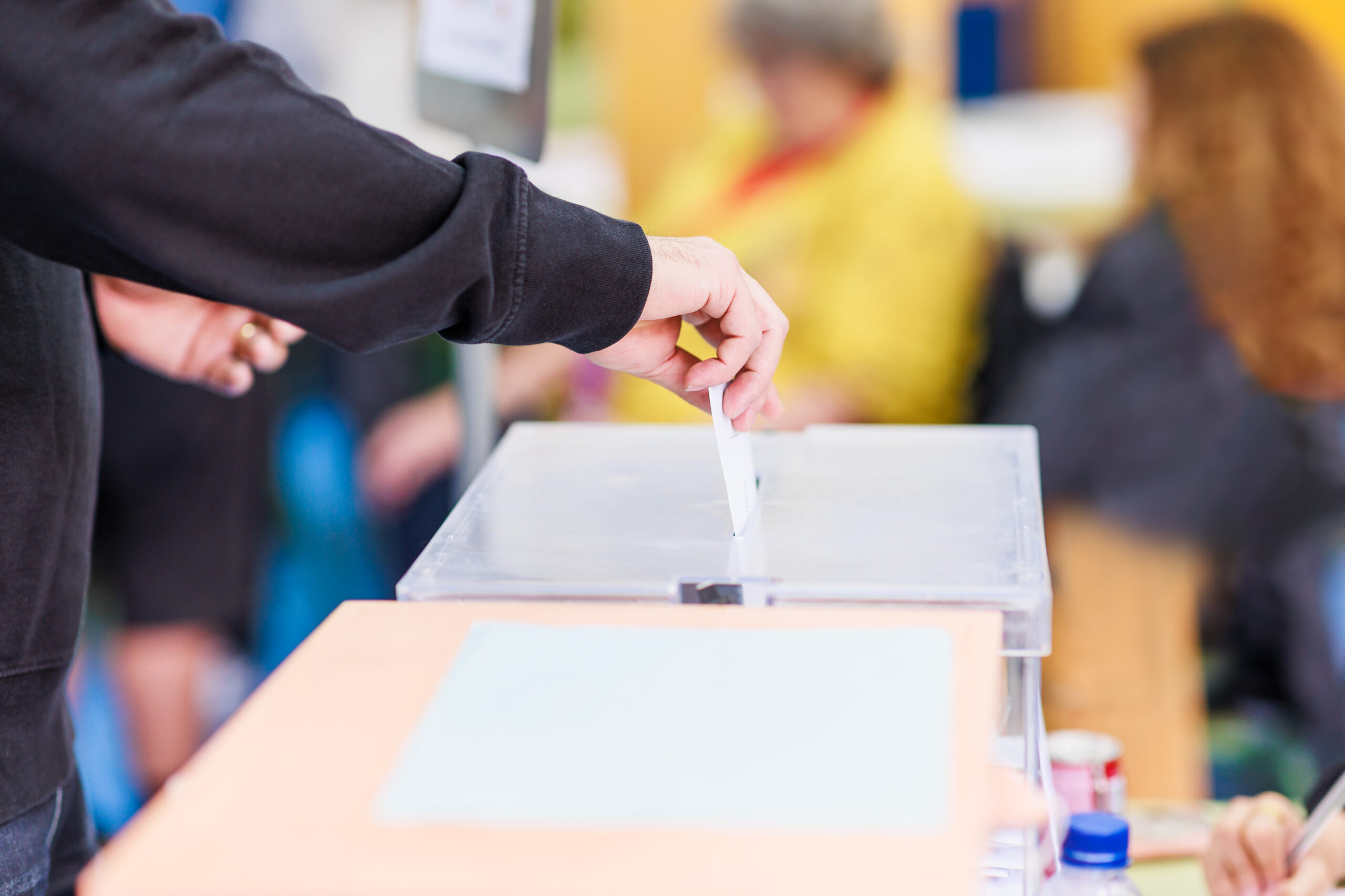 Im Vordergrund werden zwei Wahlurnen von der Seite gezeigt. Eine Person wirft einen verschlossenen Wahlumschlag in eine der Wahlurnen ein. Von der Person ist nur der Arm zu sehen.